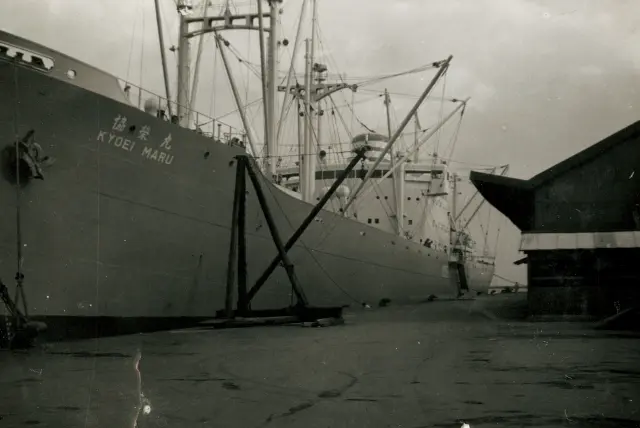 設立初年に炭酸ガス消火設備が納入された貨物船「協栄丸（KYŌEI MARU）」。岸壁から見上げるアングルで、船体と多数のデリックやマスト、右側に岸の倉庫らしき建物が見える写真。