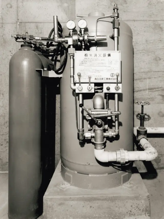 販売開始された初期の粉末消火設備。大きな縦型の消火薬剤容器と、それに隣接する小さな加圧用ガスボンベが、多数の配管、バルブ、圧力計と共にコンクリート台座の上に設置されている。