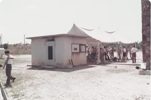 PR実験会場の外観。簡素な小屋と、その脇に設けられた大きなテントの下に多くの参加者や関係者が集まっている様子。