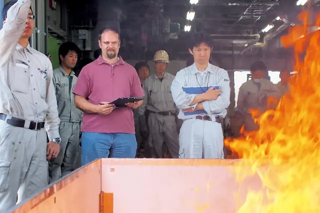UL規格追加認証試験の様子。大きな炎が上がる燃焼台の手前で、作業服姿の技術者やULの関係者が真剣な表情で消火実験を観察している。